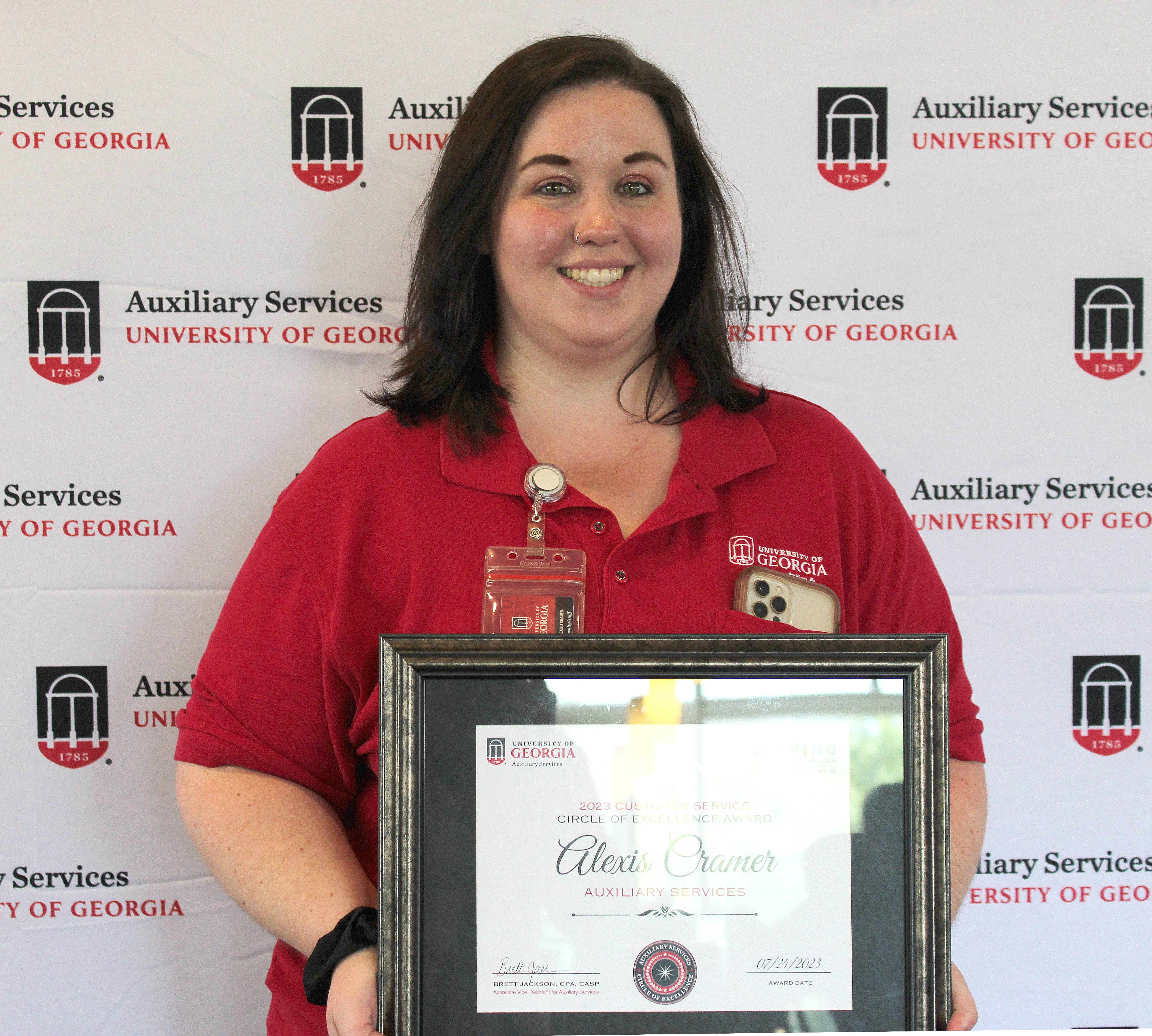 A photograph of a woman with shoulder-length brown hair wearing a red polo shirt and holding a framed certificate.