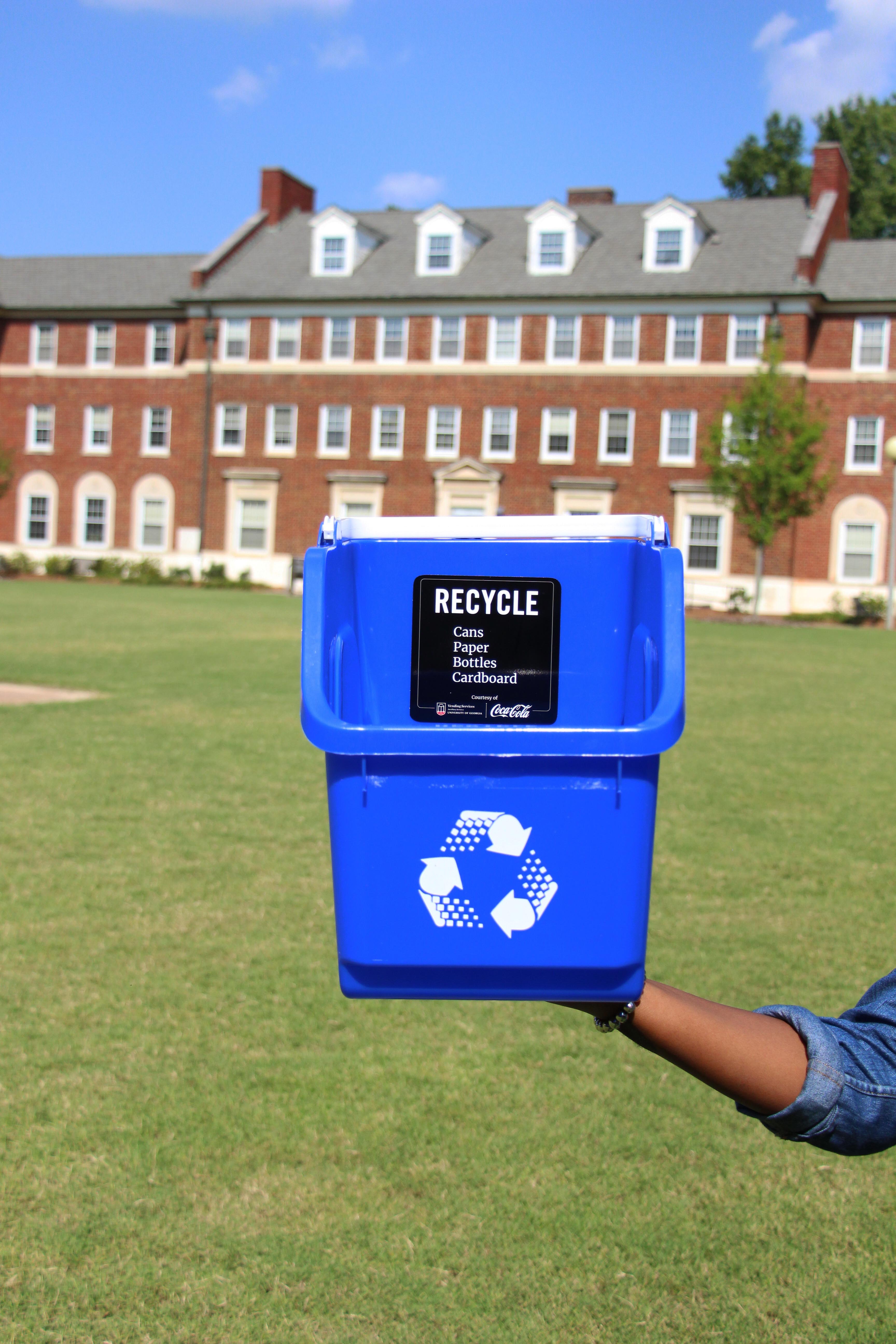 Photo of hand holding up a blue recycle bin in front of a dorm.