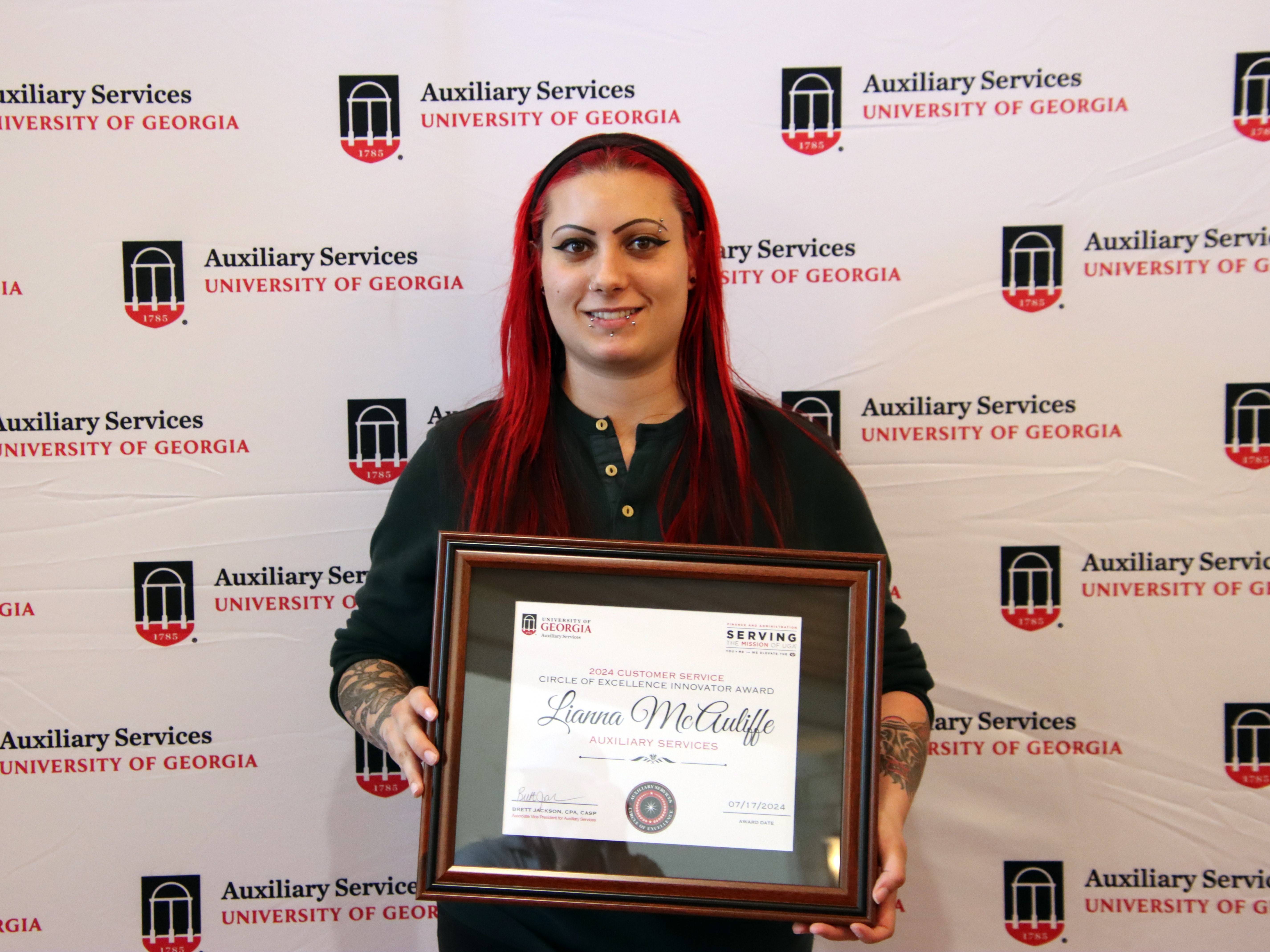 A photograph of a woman with short brown hair wearing a red polo shirt and holding a framed certificate.