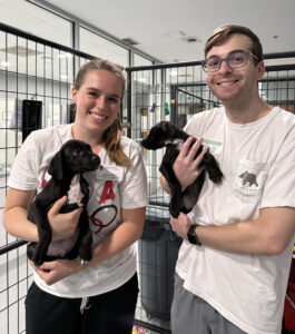 Photograph of two Auxiliary Services employees smiling and holding black puppies inside an animal shelter with metal cages in the background. Both individuals wear white shirts and name tags.