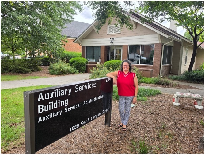 Danielle Bouton stands next to the Auxiliary Services Building sign outside of the Auxiliary Services Building. She wears a red shirt and black and white pants.
