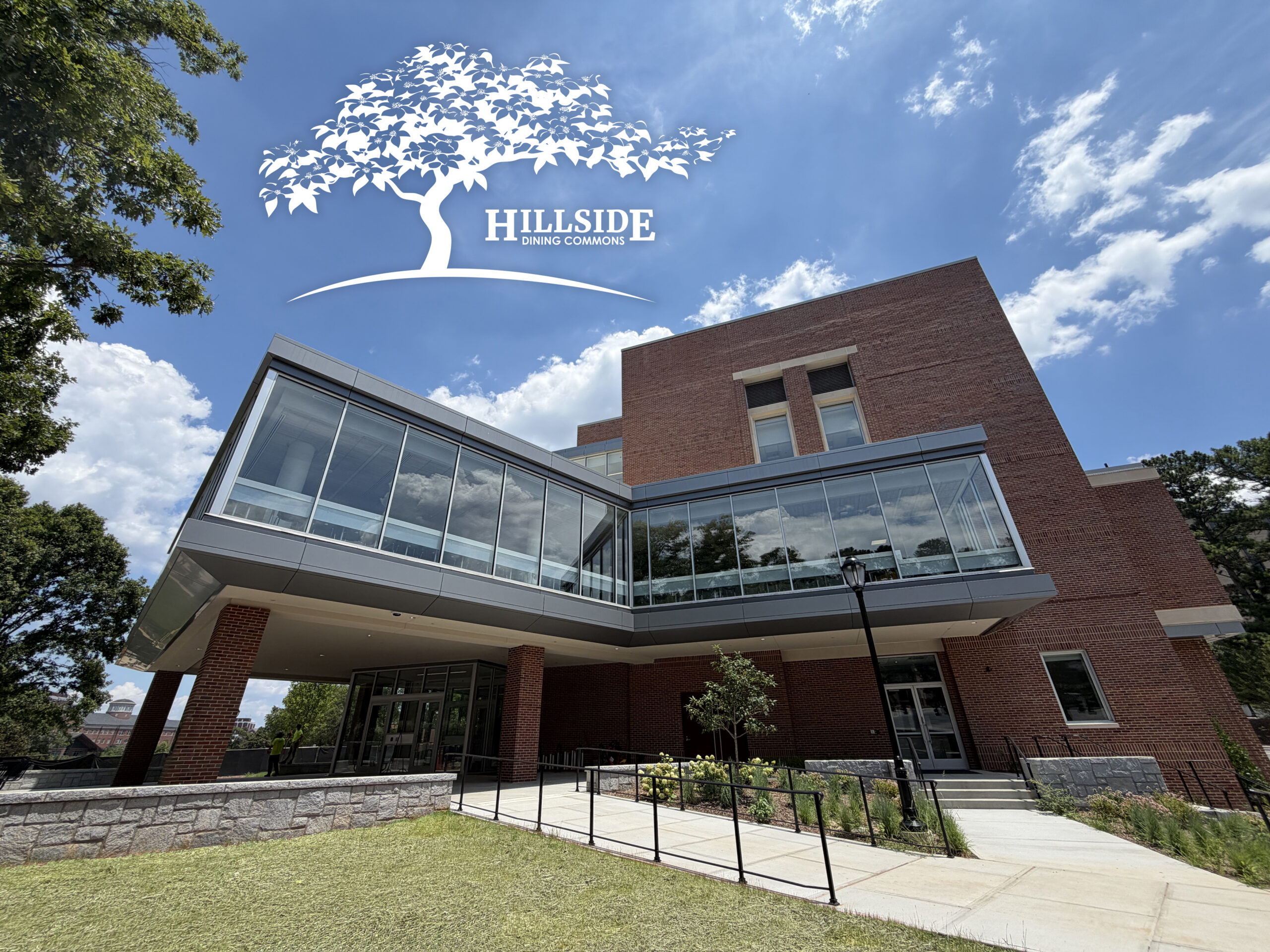 Exterior view of a rectangular brick and glass building, a blue sky with clouds, green landscaping, and the Hillside Dining Commons logo.