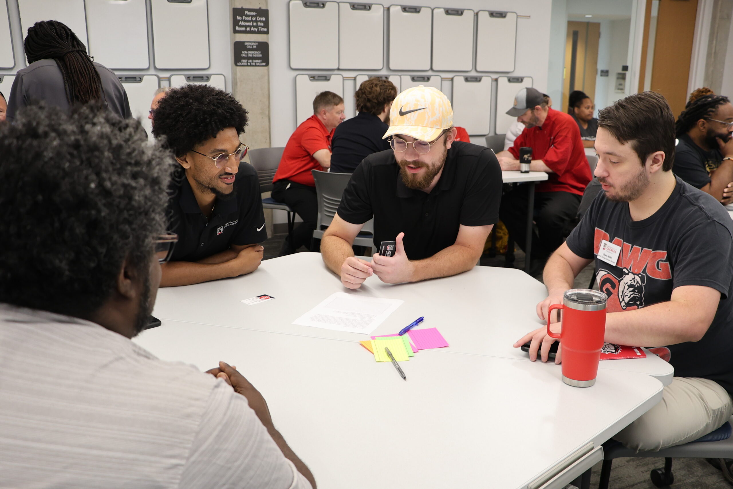 Four men are sitting at a table in a classroom filled with other individuals sitting at tables. The men are wearing short-sleeved shirts and engaged in a discussion with each other.